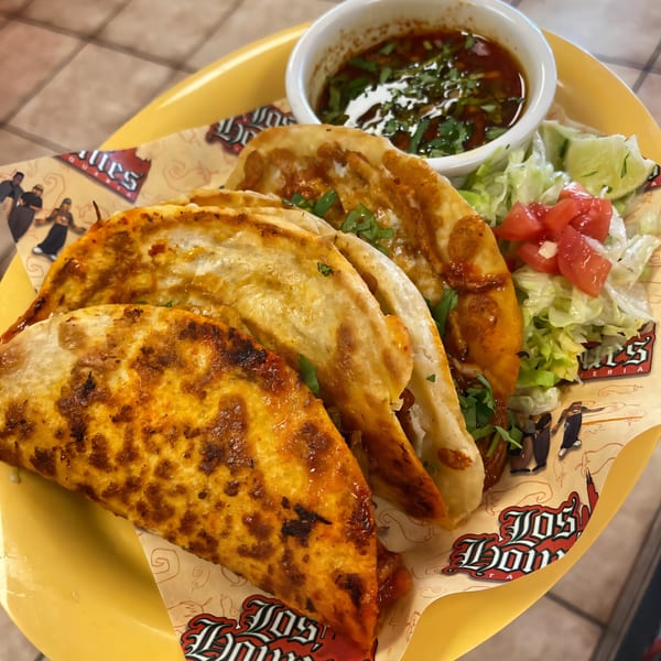 Three Los Homies Taqueria birria tacos with consommé, lettuce, and diced tomatoes on a yellow plate.